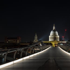 May include: A night-time photograph of St Paul's Cathedral in London, viewed from the Millennium Bridge. The cathedral's dome is lit up, with the bridge's lights leading the eye. Cranes with red lights are visible in the background.