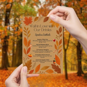 May include: A fall-themed cocktail menu held by a person. The menu features a brown background with autumn leaves and the text "Fall in Love with Our Drinks." Signature cocktails include Apple of My Eye, Spiced Maple Mule, Cinnamon Sangria, Pumpkin Patch Punch, and Caramel Apple Cider.