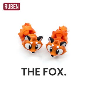 May include: A pair of orange fox-shaped earrings with black and white features. The earrings have a 3D design with a fluffy tail. The text "THE FOX." is below the earrings. The brand name "RUBEN" is in a red rectangle in the top left corner.