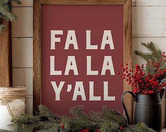 May include: A framed sign with the text "FA LA LA LA Y'ALL" in white on a burgundy background. The sign is displayed on a wooden surface, decorated with greenery, red berries, and candles, creating a festive holiday scene.