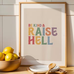 Könnte beinhalten: Ein gerahmter Druck mit dem Text "BE KIND & RAISE HELL" in bunten Buchstaben. Eine Holzschale mit Zitronen, weiße Teller und ein Holzlöffel stehen auf einer Holztheke. Der Hintergrund ist eine hellfarbige Wand.