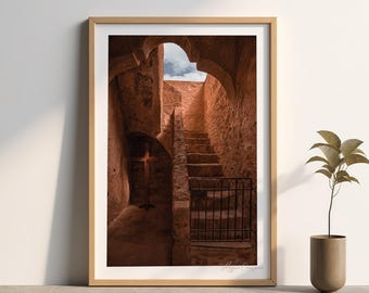 May include: Framed print depicting a stone passageway with a staircase. The walls are a warm, reddish-brown colour, and a glimpse of blue sky is visible at the top. A decorative plant in a cylindrical pot sits to the right.