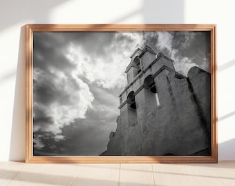 May include: Monochrome photograph of a church with a bell tower, presented in a light brown wooden frame. The sky is filled with dark, dramatic clouds. The church has a simple, classic design.