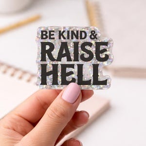 May include: A holographic sticker with the text "BE KIND & RAISE HELL" in black letters. The sticker is held by a hand with pink nail polish. The background is blurred, showing a notebook and a pen.