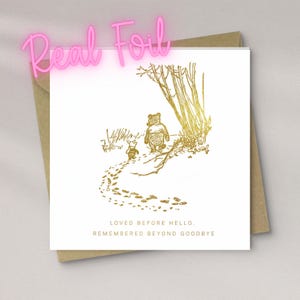 May include: A greeting card with a gold foil illustration of Winnie the Pooh and Piglet in a forest scene. The card has the text "LOVED BEFORE HELLO. REMEMBERED BEYOND GOODBYE" and is on a white background with a brown envelope.