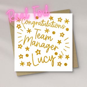 May include: A square white greeting card with gold foil text that reads Congratulations Team Manager Lucy. The card is decorated with gold foil stars and dots scattered around the text. A brown envelope is positioned behind the card.