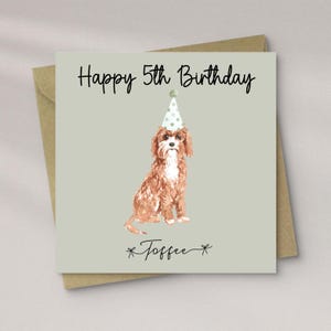 May include: A birthday card with a light grey background. It features a watercolour illustration of a Cavalier King Charles Spaniel wearing a party hat. The text "Happy 5th Birthday" is above the dog, and the name "Toffee" is below.