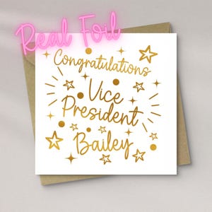May include: A square white greeting card with gold foil text that reads Congratulations Vice President Bailey. The design features gold foil stars and dots scattered around the text. The card is placed on top of a brown paper envelope.