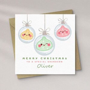 May include: A square Christmas greeting card featuring three clear glass ornaments hanging from silver strings. Inside the ornaments are glittery, smiling fig characters in gold, green, and pink. Text reads: MERRY CHRISTMAS TO A SPECIAL GRANDSON Oliver.