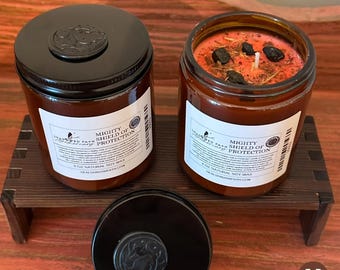 May include: Two amber glass jars with black lids and labels that read "Mighty Shield of Protection" and "8 oz Natural Soy Wax". The jars are filled with a red wax and black herbs.