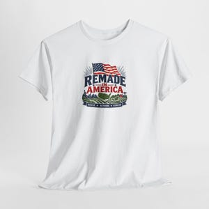 May include: A white short-sleeved t-shirt featuring a graphic print with a waving American flag above the text REMADE IN AMERICA. Below the text is a landscape illustration of a city skyline, a map of the USA, and fields with the text REUSE RETHINK RENEW.