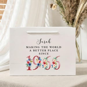 Op de afbeelding: Witte cadeauzak met de naam "Sarah" in cursief schrift. De tekst "MAKING THE WORLD A BETTER PLACE SINCE 1965" is eronder gedrukt. De cijfers in "1965" zijn versierd met bloemmotieven.