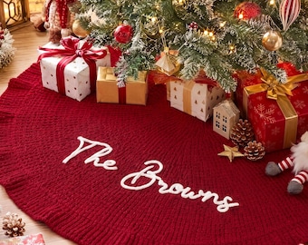 Peut inclure: Une jupe de sapin de Noël tricotée en forme circulaire rouge foncé avec un bord volanté et le texte brodé en blanc "The Browns" en écriture cursive. La jupe est placée sous un sapin de Noël décoré avec des cadeaux et une figurine de gnome.