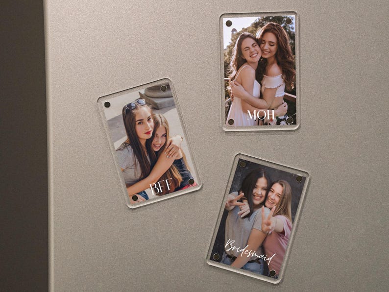 May include: Three rectangular clear acrylic magnets with photos of people. One magnet has the word "BFF", another has "MOH", and the third has "Bridesmaid" written on them. The magnets are on a gray surface.