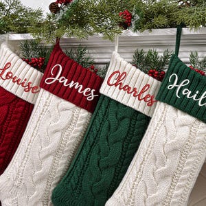 May include: Four cable-knit Christmas stockings in red, cream, and green, each with a contrasting cuff embroidered with a name in white script: Louise, James, Charles, and Hailey. The stockings are hanging on a white mantelpiece, decorated with greenery and red berries.
