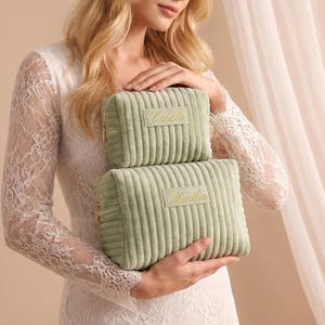 May include: Two sage green corduroy cosmetic bags, one stacked on top of the other. The top bag has the name "Calista" embroidered in gold, and the bottom bag has "Martha" embroidered in gold. Both bags have gold zippers.