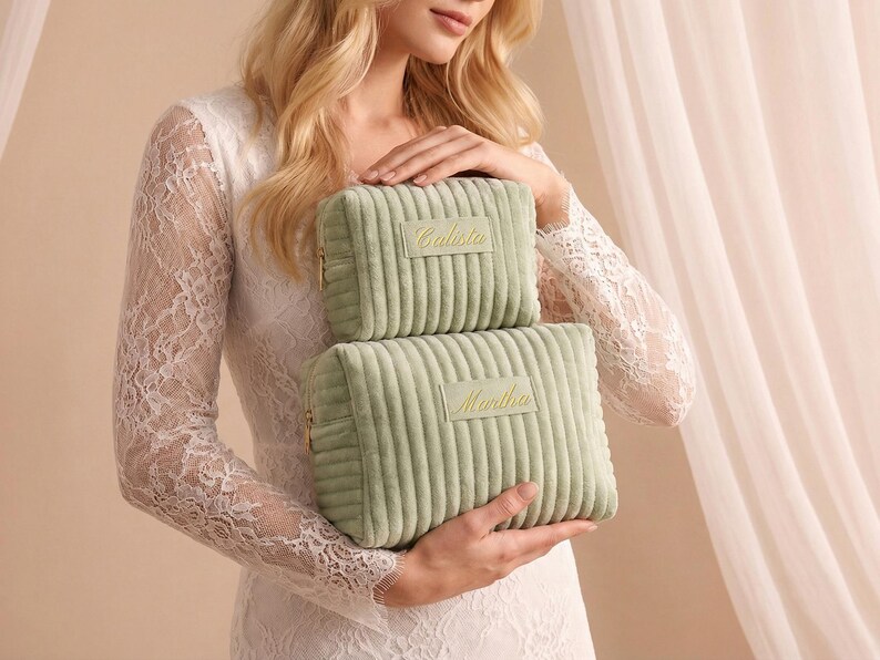 May include: Two sage green corduroy cosmetic bags, one stacked on top of the other. The top bag has the name "Calista" embroidered in gold, and the bottom bag has "Martha" embroidered in gold. Both bags have gold zippers.