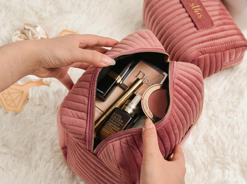May include: A dusty rose quilted velvet cosmetic bag, open to reveal makeup products. The bag is filled with items like a lipstick, eyeshadow, and a bottle of Estee Lauder Advanced Night Repair. Another bag is in the background.