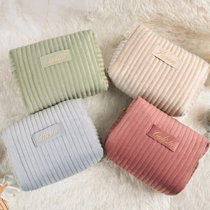 May include: Four corduroy cosmetic bags in pastel colors: sage green, light pink, light blue, and beige. Each bag has a rectangular label with a name embroidered in gold. The bags are arranged on a fluffy white surface.