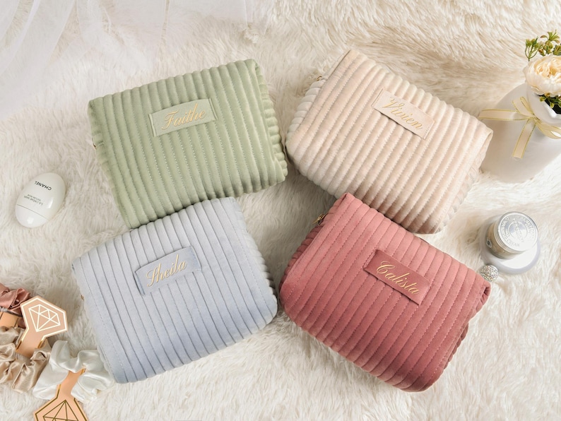 May include: Four corduroy cosmetic bags in pastel colors: sage green, light pink, light blue, and beige. Each bag has a rectangular label with a name embroidered in gold. The bags are arranged on a fluffy white surface.