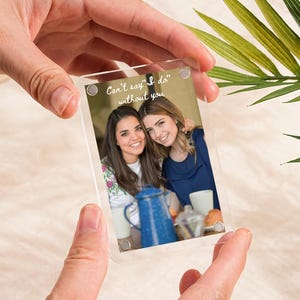 May include: A clear acrylic photo frame with a photo of two smiling women. The photo includes a blue enamelware coffee pot and the text "Can't say 'I do' without you". The frame is held by two hands.