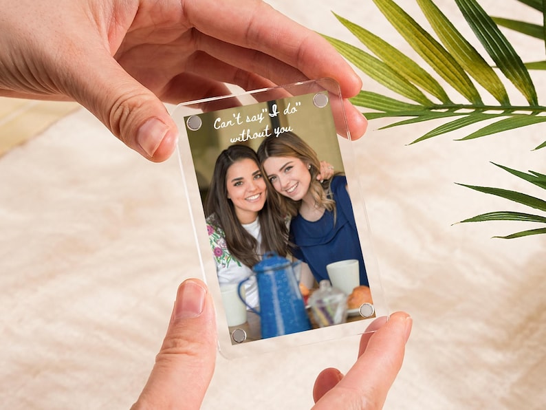 May include: A clear acrylic photo frame with a photo of two smiling women. The photo includes a blue enamelware coffee pot and the text "Can't say 'I do' without you". The frame is held by two hands.