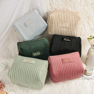 May include: A collection of corduroy cosmetic bags in various colors, including light blue, beige, green, black, light green, and pink. Each bag features a rectangular label with a name in gold lettering and a gold zipper.