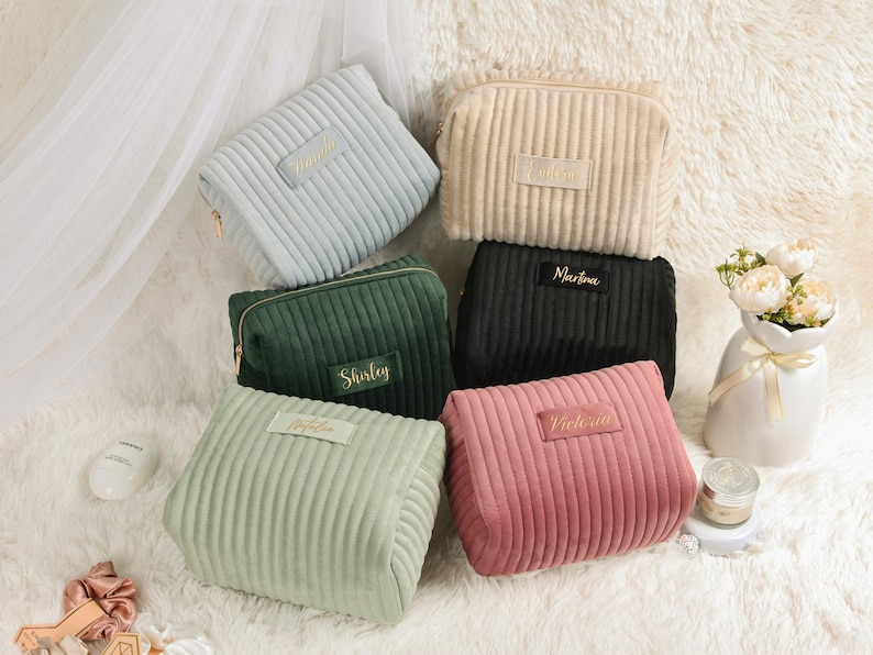 May include: A collection of corduroy cosmetic bags in various colors, including light blue, beige, green, black, light green, and pink. Each bag features a rectangular label with a name in gold lettering and a gold zipper.