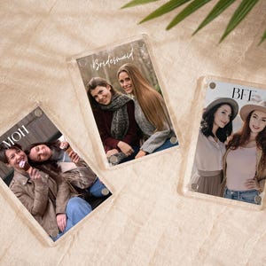 May include: Three clear acrylic photo frames with pictures of people. One frame says "MOH", another says "Bridesmaid", and the third says "BFF". The frames are on a beige surface with green leaves in the background.