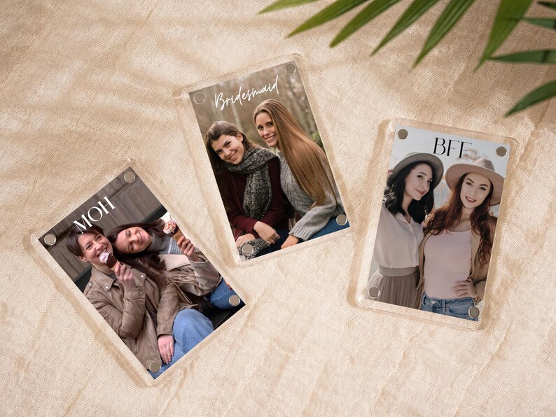 May include: Three clear acrylic photo frames with pictures of people. One frame says "MOH", another says "Bridesmaid", and the third says "BFF". The frames are on a beige surface with green leaves in the background.