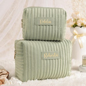 May include: Two sage green corduroy cosmetic bags stacked on top of each other. The top bag has the name "Odelia" in gold lettering, and the bottom bag has the name "Roberta". Both bags have a zipper closure.