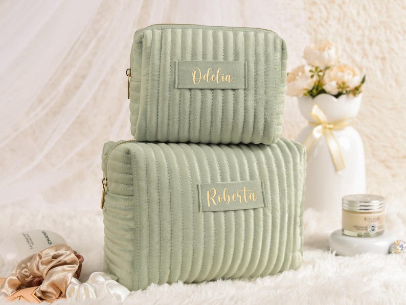 May include: Two sage green corduroy cosmetic bags stacked on top of each other. The top bag has the name "Odelia" in gold lettering, and the bottom bag has the name "Roberta". Both bags have a zipper closure.