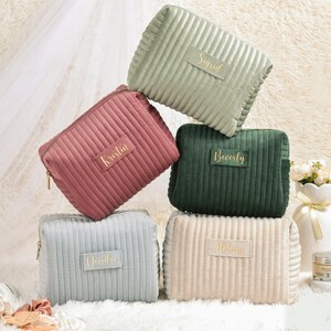 May include: A stack of five corduroy cosmetic bags in various colors: sage green, dusty rose, forest green, light blue, and cream. Each bag has a gold zipper and a gold nameplate with a name in gold lettering. The bags are arranged on a fluffy white surface.