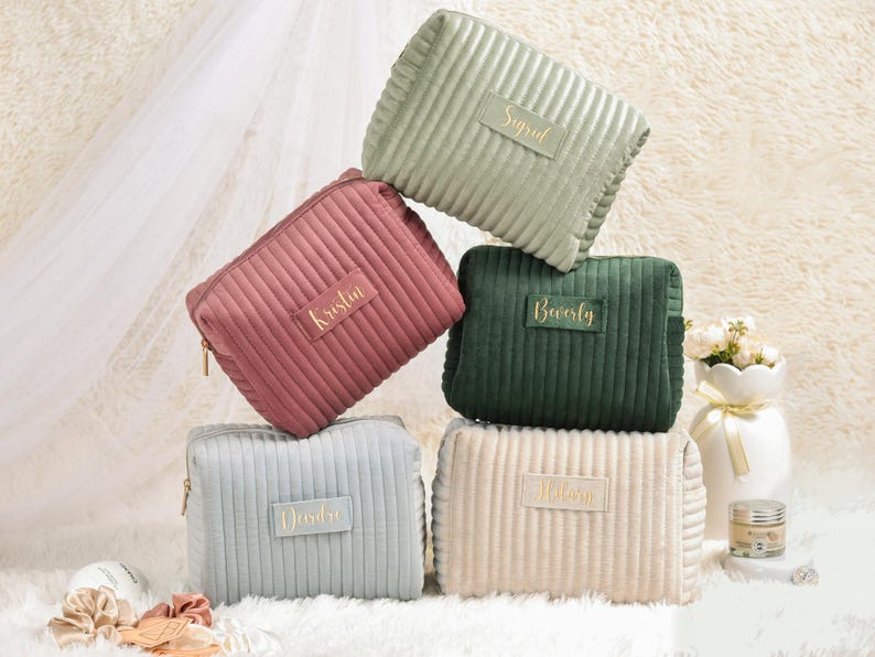 May include: A stack of five corduroy cosmetic bags in various colors: sage green, dusty rose, forest green, light blue, and cream. Each bag has a gold zipper and a gold nameplate with a name in gold lettering. The bags are arranged on a fluffy white surface.