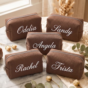 May include: Five rectangular brown waffle-textured cosmetic bags with silver zips. Each bag features a white name printed in a cursive script: Odelia, Sandy, Angela, Rachel, and Trista. The bags are arranged in a group on a marble surface.