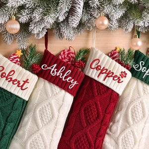 May include: Four personalized Christmas stockings in green, white, and red, with names embroidered in cursive. The stockings are knit with cable and diamond patterns, and decorated with festive greenery and ornaments.