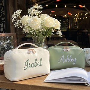 May include: Two rectangular cosmetic bags with top handles and rose gold zipper pulls. The white bag features the name Isabel embroidered in green, while the sage green bag features the name Ashley printed in white script. Both have smooth finishes.