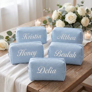 May include: Five light blue rectangular cosmetic bags with a waffle-textured fabric. Each bag features a white embroidered name on the front, specifically the names Kristin, Althea, Honey, Beulah, and Delia, with a silver zipper closure on the side.