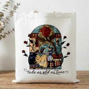 Puede incluir: Bolso tote blanco con un diseño de vidriera con una rosa roja y una pareja bailando. Las palabras "tale as old as time" están escritas en una fuente cursiva. La bolsa está hecha de tela resistente.