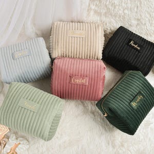 May include: A collection of six corduroy cosmetic bags in various colors: light blue, beige, pink, black, and green. Each bag has a rectangular shape with a zipper closure and a label with a name in gold lettering. The bags are arranged on a white, fluffy surface.