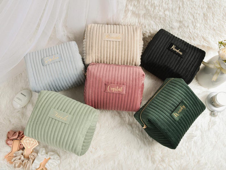 May include: A collection of six corduroy cosmetic bags in various colors: light blue, beige, pink, black, and green. Each bag has a rectangular shape with a zipper closure and a label with a name in gold lettering. The bags are arranged on a white, fluffy surface.