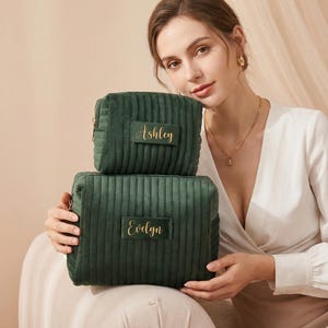 May include: Two dark green, corduroy cosmetic bags, one stacked on top of the other. The top bag has the name "Ashley" in gold script, and the bottom bag has "Evelyn". The bags are rectangular with a zipper closure.