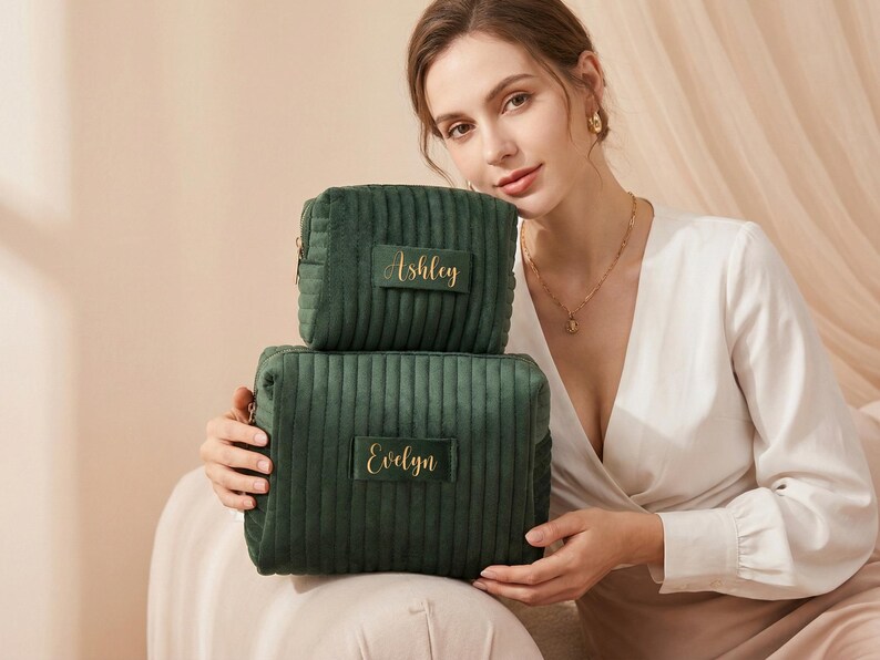 May include: Two dark green, corduroy cosmetic bags, one stacked on top of the other. The top bag has the name "Ashley" in gold script, and the bottom bag has "Evelyn". The bags are rectangular with a zipper closure.