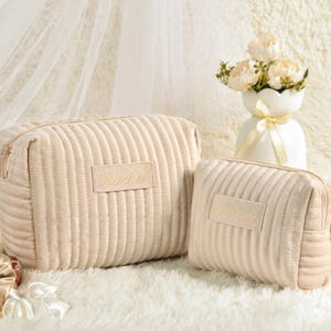 May include: Two beige corduroy cosmetic bags with gold zippers and the name "Rachel" embroidered on the front. The bags are on a white fluffy surface, with a vase of flowers in the background.