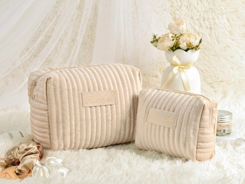 May include: Two beige corduroy cosmetic bags with gold zippers and the name "Rachel" embroidered on the front. The bags are on a white fluffy surface, with a vase of flowers in the background.