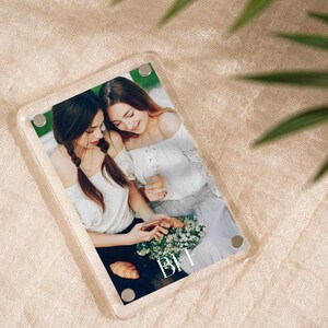 May include: A clear acrylic photo frame with a picture of two women. The women are wearing white tops and are holding flowers and drinks. The letters "BFF" are printed on the photo. The frame has four silver magnetic closures.