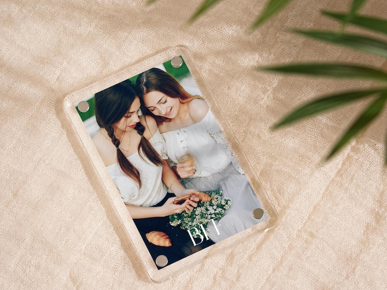 May include: A clear acrylic photo frame with a picture of two women. The women are wearing white tops and are holding flowers and drinks. The letters "BFF" are printed on the photo. The frame has four silver magnetic closures.