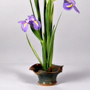 May include: Purple iris flowers arranged in a green and brown ceramic vase. The vase has a unique, wavy shape.