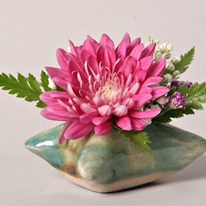May include: A pink chrysanthemum flower arrangement in a green ceramic vase. The vase is shaped like a pillow and has a textured surface.