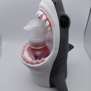 May include: A shark-shaped soap dispenser in gray, white, and black. The shark's mouth is open, revealing white teeth and a pink interior. A clear soap bottle with a pink liquid is inside the shark's mouth. The dispenser has a whimsical design.